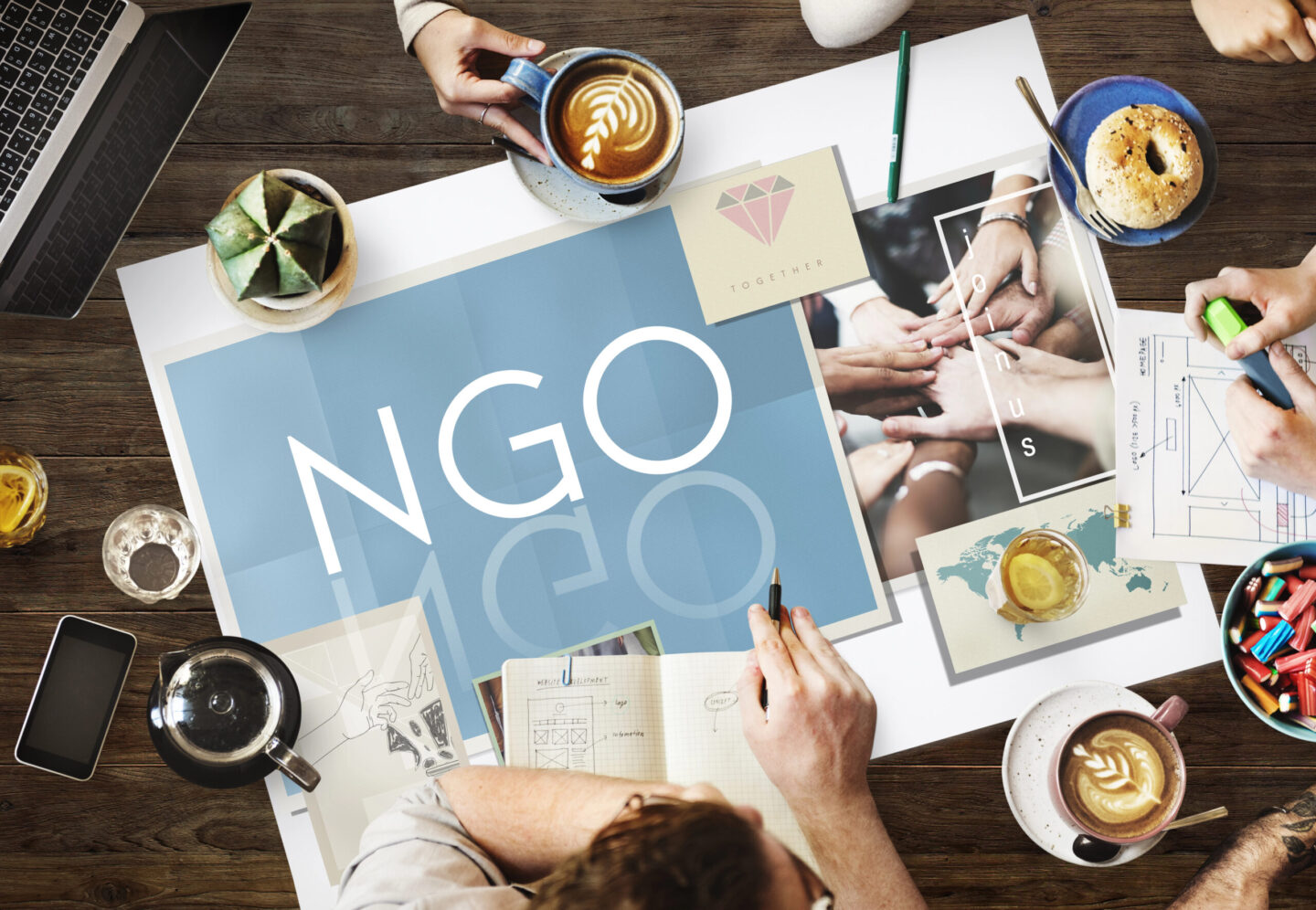 Grupa ludzi omawiająca projekty NGO przy stole, na którym znajdują się dokumenty, notatki i napoje, z napisem 'NGO' w centralnym miejscu.